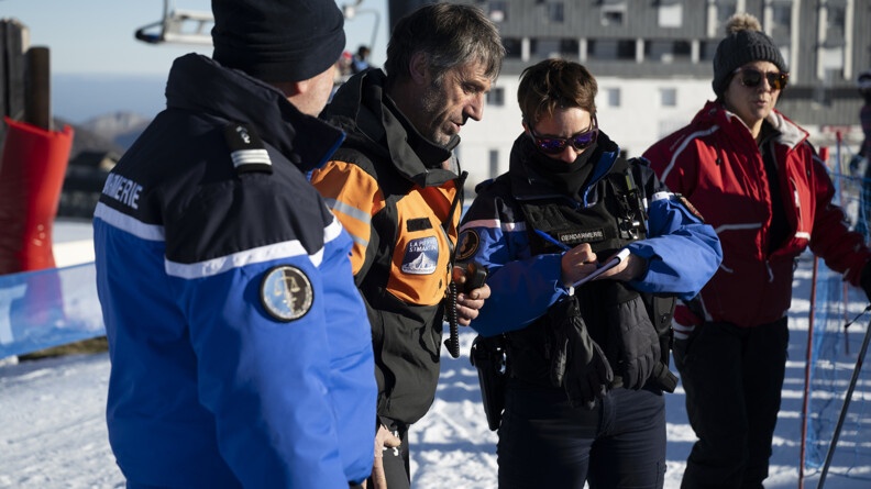 Deux gendarmes dont une femme prenant des notes, avec un homme en tenue orange et noir du nom de la ville, au bord des pistes