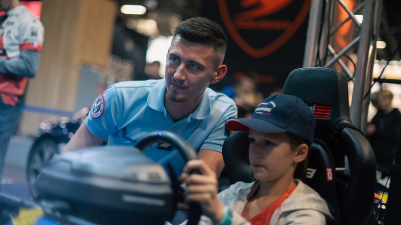 Un gendarme en tenue est accroupi aux côtés d'un jeune garçon au volant d'un simulateur de conduite. Le garçonnet porte une casquette bleue marine sur laquelle on devine l'inscription Gendarmerie.