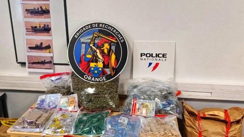 Agrandir l'image Sur une table, de nombreux sacs de scellés transparents contenant du cannabis, de l'argent liquide, des contenants, surmontés des rondaches de la brigade de recherches d'Orange et de la police nationale. De part et d'autre de la table et devant elle, de nombruex sacs krafts de scellés. Au mur, accroché à un tableau, cinq photos d'armes longues