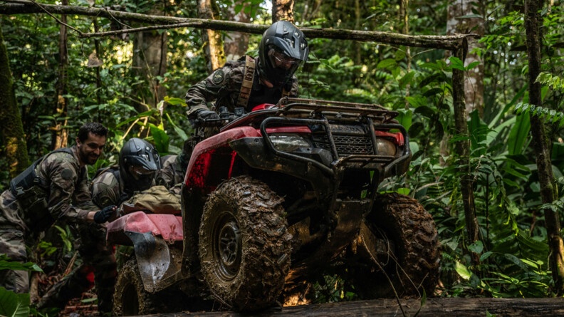 Militaire faisant passer son quad au dessus d'un tronc avec l'aide de deux camarades.