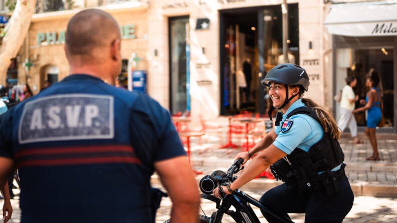 Une femme gendarme, en vélo, dans une rue passante, regarde en souriant un agent portant dans le dos la mention "A.S.V.P."