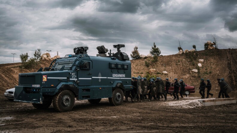 Un peloton de gendarmerie mobile en colonne d'assaut derrière un Centaure sur un terrain boueux. Derrière eux, un talus