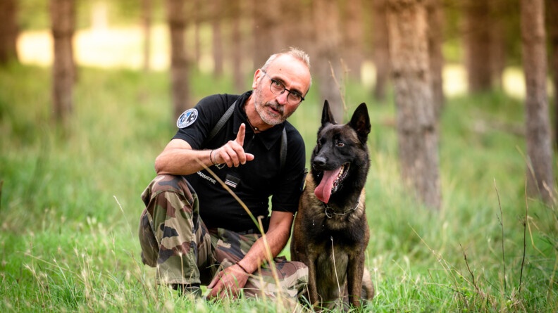 Maître chien Major Yves accroupi dans l'herbe et son chien Tirock positionné à ses côtés. Le maître chien pointe son doigt vers quelque chose et semble donner ses consignes à son chien qui se montre à l'écoute.