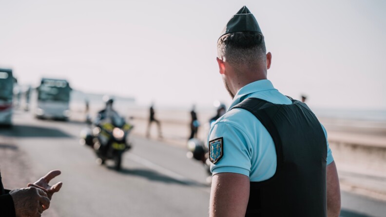 Un gendarme de dos en polo bleu avec dans le fond en flou, un motocycliste de la gendarmerie nationale.