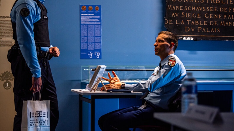 Le colonel R&eacute;my Nollet &eacute;change avec un sous lieutenant, nous voyons la vue de c&ocirc;t&eacute; avec sa table et son pr&eacute;sentoir avec son livre dessus.