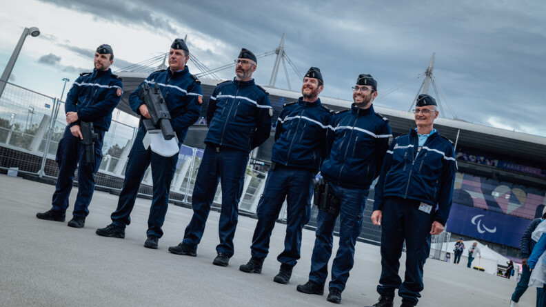Six gendarmes se tiennent debout, côte à côte, devant le stade de France (Saint-Denis). L'un d'eux tient dans ses mains un fusil anti-drone.