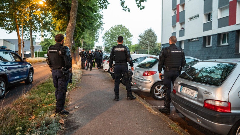 de dos, trois gendarmes en tenue noire et gilet pare-balles, derrière un attroupement et leur collègues, à droite des véhicules garés en bas de barres d'immeubles