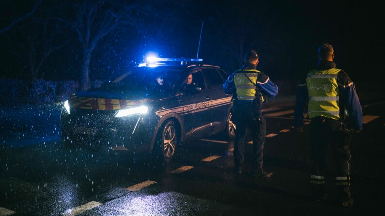 4 gendarmes de nuit sont sur la route pour un contrôle routier. Sur la droite, 2 d'entre eux sont debout de dos. Et sur la gauche, 2 autres sont dans un véhicule, phares allumés, sous la pluie.