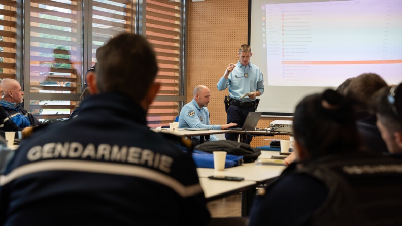 Dans une salle se tient une réunion. En premier plan, on voit un gendarme assis de dos portant une veste bleue marine portant la mention "Gendarmerie". A ses côtés, on aperçoit d'autres gendarmes, hommes et femmes, également assis, de dos ou de biais. devant eux, un gendarme, homme d'âge moyen, se tient debout, en train de parler. Derrière lui, un grand écran avec une projection sur le mur.