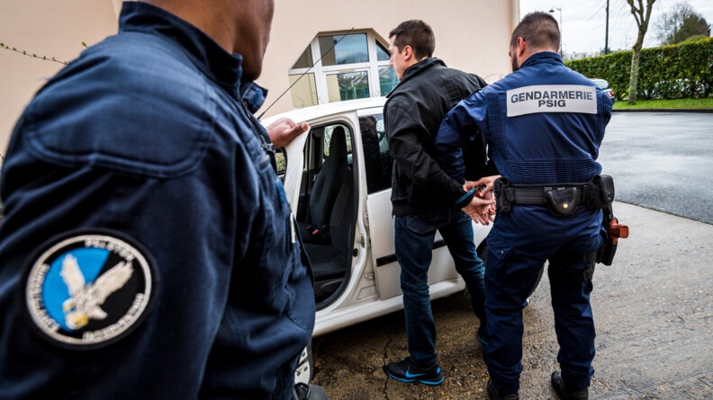 Deux gendarmes en combinaison bleue-marine, un en train de tenir une portière de voiture à gauche et l'autre en train de passer les menottes à un individu devant une voiture blanche. Au fond un bâtiment et à droite la route, une haie, un arbre