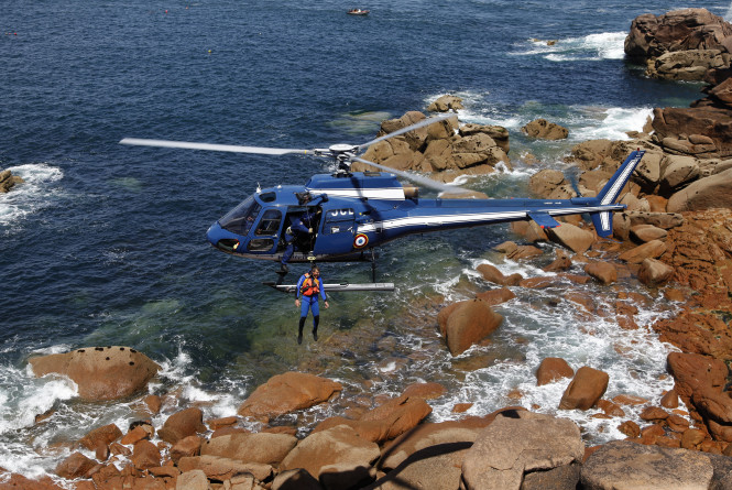 Un h&eacute;licopt&egrave;re bleu marine de la Gendarmerie nationale est en vol stationnaire au-dessus d&rsquo;un littoral rocheux. Un plongeur en tenue de sauvetage est suspendu sous la porte lat&eacute;rale, au dessus de l'eau. Le paysage est constitu&eacute; de gros rochers arrondis, partiellement immerg&eacute;s, et d&rsquo;une mer bleue et calme.