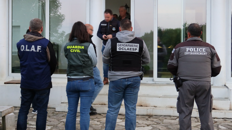 Un agent de l'OLAF, une enqu&ecirc;trice de la Guardia civil, un enqu&ecirc;teur de l'OCLAESP et un agent de l'OFB, en civil, avec une chasuble aux couleurs de leur unit&eacute; d'appartenance, vus de dos, devant l'entr&eacute;e d'un b&acirc;timent dont sort un autre gendarme de l'OCLAESP.
