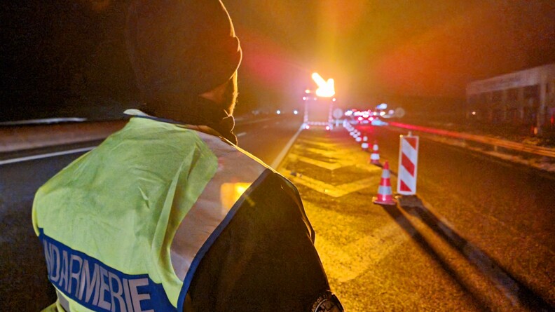 photo de nuit, avec des éclairages lumineux orangés. Vue de dos sur un gendarme de l'escadron de sécurité routière du GGd28, portant une chasuble jaune fluo floquée gendarmerie. Il regarde vers l'avant, où est déployé un véhicule à flècle lumineuse porté vers la droite.