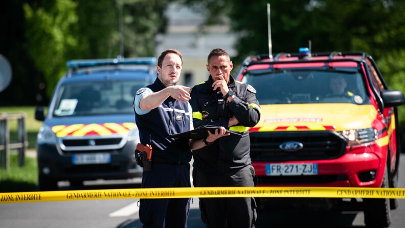 Un gendarme discute avec un personnel de la sécurité civile et désigne un objet. Les deux se trouvent derrière une rubalise. Deux véhicules se trouvent en arrière plan.