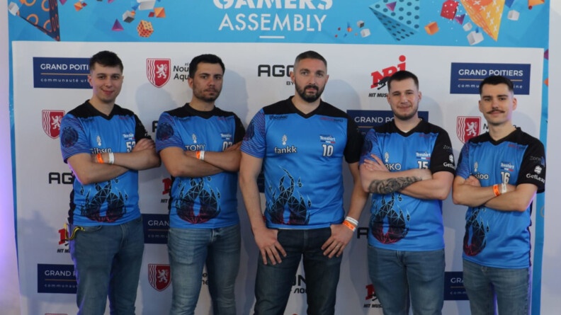 Agrandir l'image 5 hommes vêtus de jean et d'un t-shirt bleu et noir, dont toris les bras croisés, debouts devant un panneau de sponsors