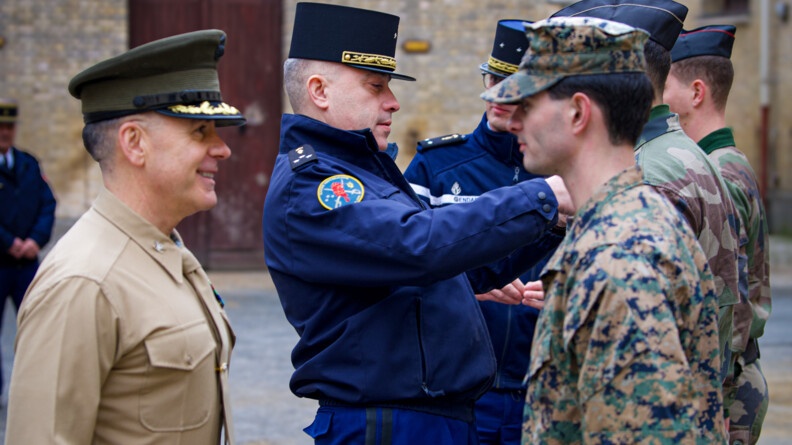 Un officier des marines éhcnage un regard avec son subordonné; en arrière-plan un général de la Garde remet un brevet à un militaire
