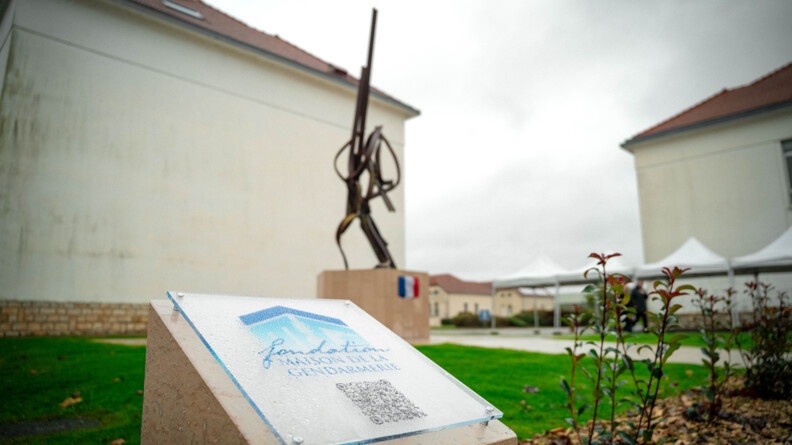 Une plaque en plexi trabsparent porte l'inscription "Fondation maison de la gendarmerie", sous laquelle apparait un QR Code. En arri&egrave;re plan, on aper&ccedil;oit une sculpture monumentale en acier, pos&eacute;e sur un socle de couleur claire, au devant duquel est accroch&eacute; un drapeau bleu blanc rouge