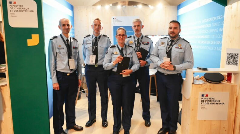 Au stand MIOM de l'ANCTour, cinq gendarmes en chemise bleue, cravate et pantalon droit, posent. A gauche, deux colonels. Au centre une adjudant-chef (ADC) référente sûreté, et son camarade ADC référent sûreté à droite, tenant tous les deux une tablette neogend. A droite, un autre adjudant-chef.
