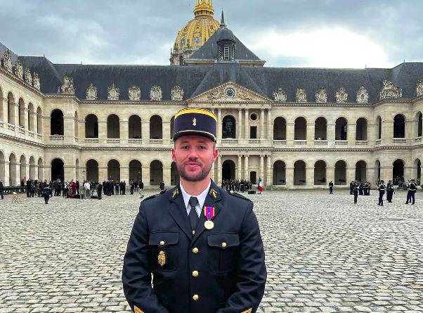 Agrandir l'image Un jeune homme brun, portant la barbe, en uniforme bleu marine et un képi bleu marine et doré, pose debout, les deux mains jointes devant lui, dans une cour pavée (il s'agit de la cour d'honneur des invalides). A l'arrière, on aperçoit le dôme des invalides, ainsi que le ciel gris.