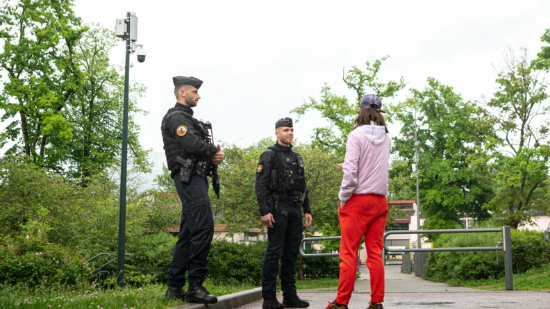 Agrandir l'image Deux gendarmes discutent avec un adolescent portant une casquette violette, un blouson rose, un jogging rouge et des baskets. lls se tiennent debout, dans l'espace extérieur et verdoyant d'une résidence