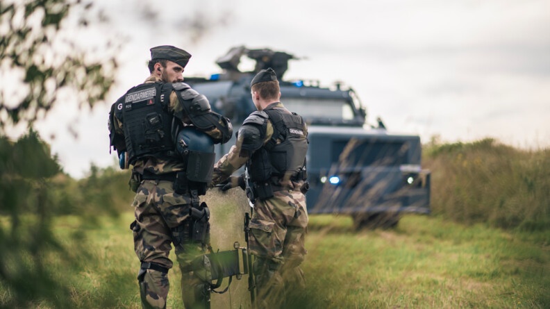 Agrandir l'image Deux gendarmes de dos au premier plan. En arrière-plan, en flou, un véhicule Centaure.