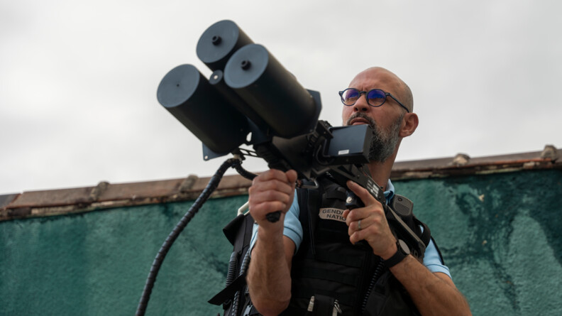 Homme de face, debout, habillé en tenue de gendarme, portant un appareil de brouillage multi-fréquences orienté vers le ciel