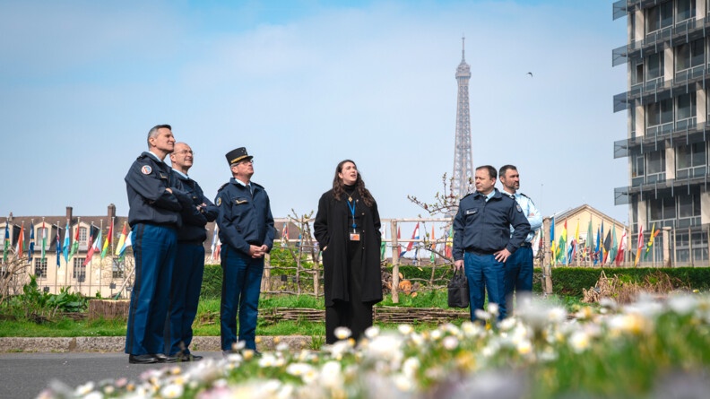 Agrandir l'image Cinq gendarmes et une femme en civil dans un jardin. EN arrière-plan à gauche plusieurs drapeaux de différents pays, en arrière-plan au fond la tour-eiffel