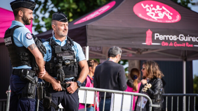 A gauche, deux gendarmes en sécurisation, devant des barrières de sécurité bordant le public. En arrière-plan à droite, une tonnelle marron en rose de la ville de Perros-Guirec