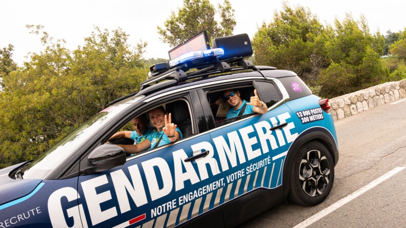 Agrandir l'image Une voiture de couleur bleue marine de la gendarmerie roule sur la route. On aperçoit les gendarmes qui sont à son bord, souriants, et effectuant le signe de la victoire avec leurs doigts, par les fenêtres ouvertes.