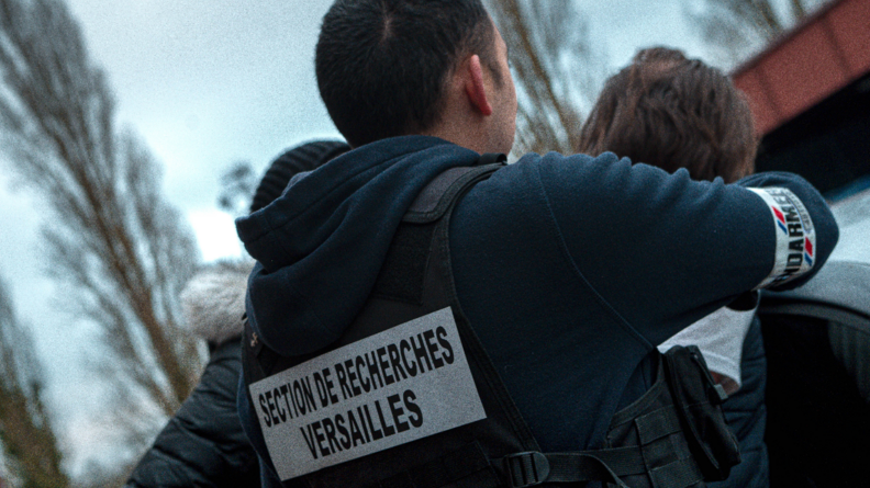 Photo d'un gendarme de dos, en train d'intercepter un suspect. Au dos de sa veste est inscrit SECTION DE RECHERCHES VERSAILLES. Il porte aussi un brassard portant l'incription GENDARMERIE.