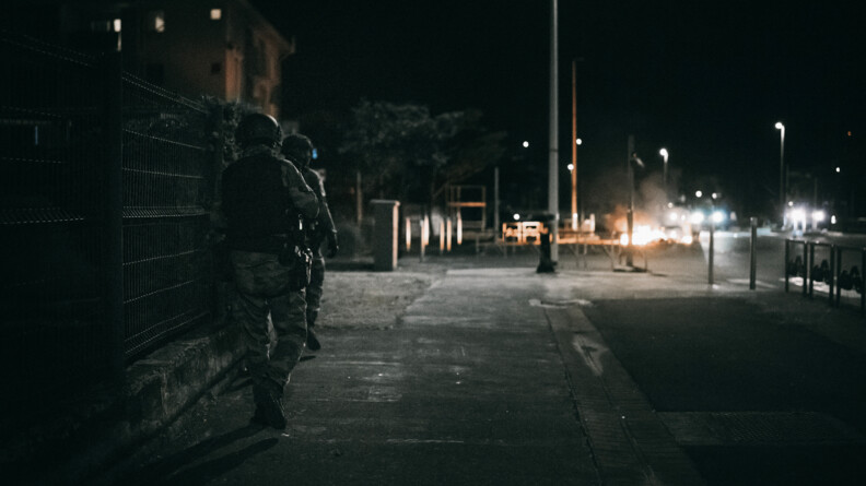 De nuit, deux gendarmes en treillis équipés de gilets lourds patrouillent le long d'une clôture. Au tour d'eux le mobileir urbain, avec uà droite la route, éclairée au fond par des pétards et feux
