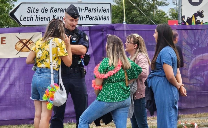 Agrandir l'image Un Un gendUn gendarme en tenue d'intervention, debout, entouré par quatre jeunes femmes. Derrière eux, un panneau de direction et une bâche violette.