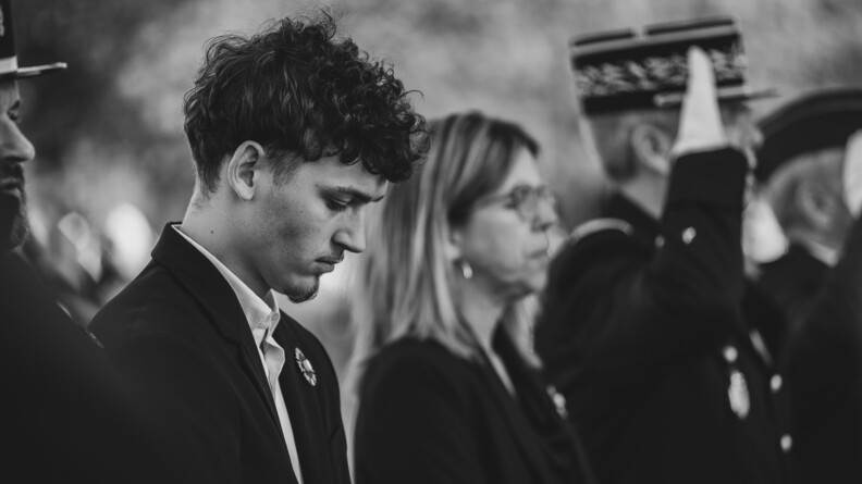 Portait de profil d'un jeune homme brun, portant une veste noire réhaussée d'un Bleuet de France. A sa droite, on devine celui d'un gendarme. A sa gauche, plus flous, une femme ferme les yeux et un général de gendarmerie salue