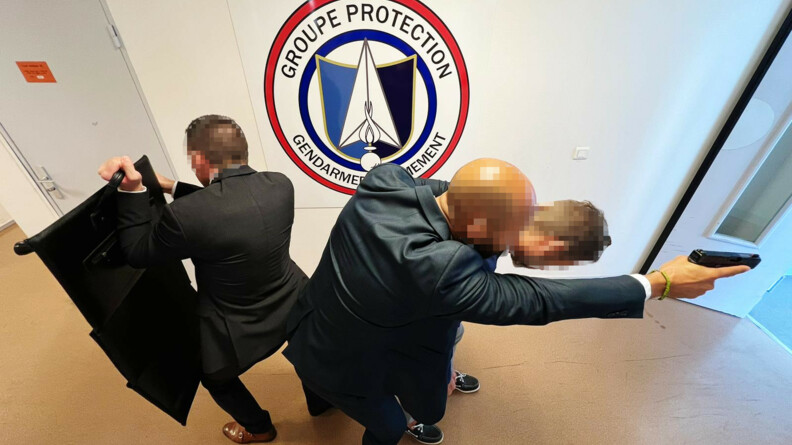 Deux hommes en costume, dos à dos, debout, dans un espace intérieur. L'un deux protège de son bras gauche une personne. Il tient un pistolet dans sa main droite. Celui qui lui tourne le dos tient déployée devant lui une mallette pare-balles. En gros sur le mur derrière est inscrit Groupe protection - Gendarmerie Armement