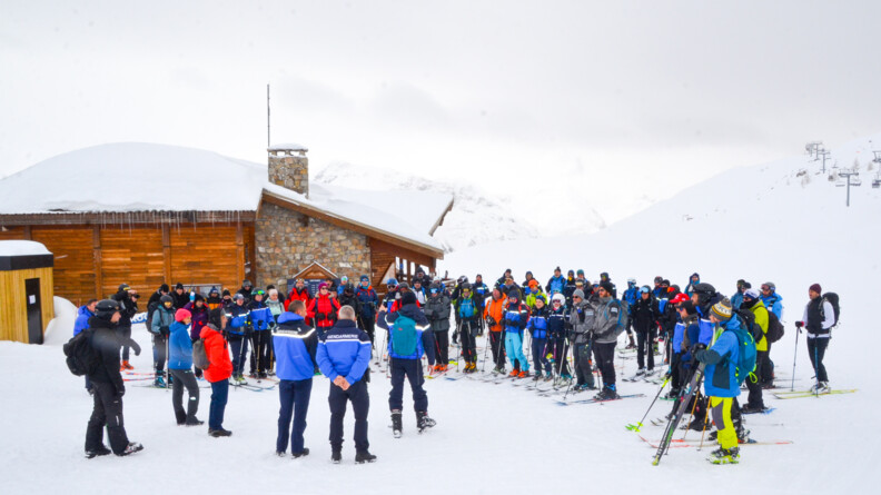 Devant un châlet sur la gauche, une soixantained personnes à skis rassemblées autour de 3 gendarmes
