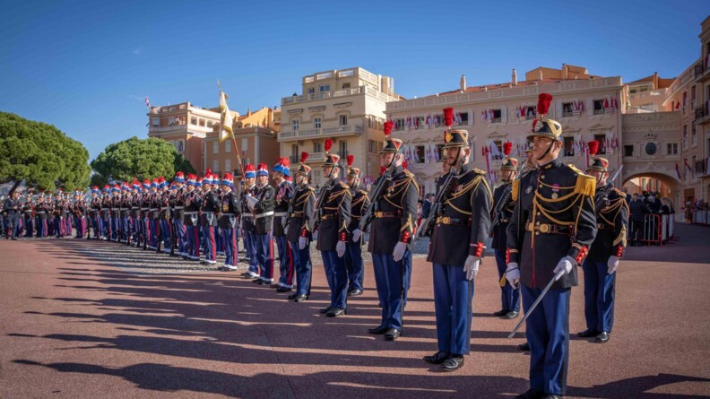 Devant des bâtiments auc couleurs claires et sur un sol rougeâtre, des drapeau blanc et rouge aux fenêtres, les militaires de la garde républicaine alignés avec ceux de la garde de Monaco