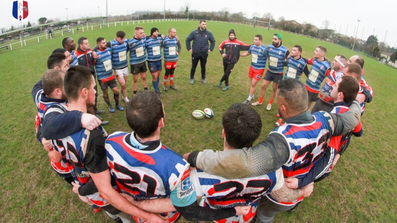 Vue en plong&eacute;e du XV gendarmerie se tenant par les &eacute;paules et le dos en cercle, dans un stade. Au centre du cercle, deux ballons de rugby