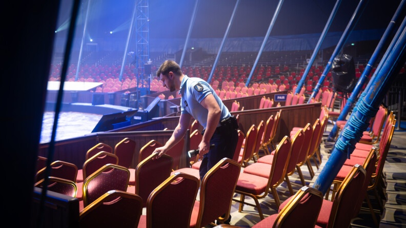 Un gendarme muni d'une lampe torche inspecte le dossier d'une chaise dans un chapiteau où sont disposées de nombreuses autres chaises autour de la scène. Le lieu est éclairé par des spots lumineux.