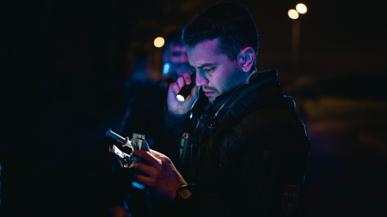 De nuit, il fait sombre, un gendarme regarde avec une lampe torche, les papiers d'un véhicule qu'il vient de contrôler.