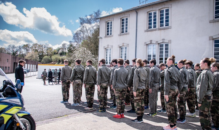 Une vingtaine de jeunes vêtus de treillis et baskets en formation ordre serré devant des bâtiments. A gauche de la photo, l'avant d'une moto gendarmerie