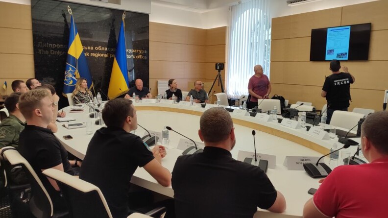 Une quinzaine de personnes assises autour d'une table blanche ovale. A gauche, devant une plaque de marbre noire, les drapeaux ukrainiens. A droite, au second plan, un expert de l'IRCGN vu de dos donne des explications devant un &eacute;cran de t&eacute;l&eacute;vision.
