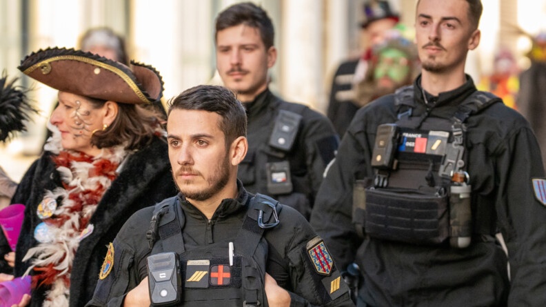 La photo représente trois gendarmes avançant dans la rue aux côtés des carnavaleux, portant un déguisement, lors du carnaval de Bergues.