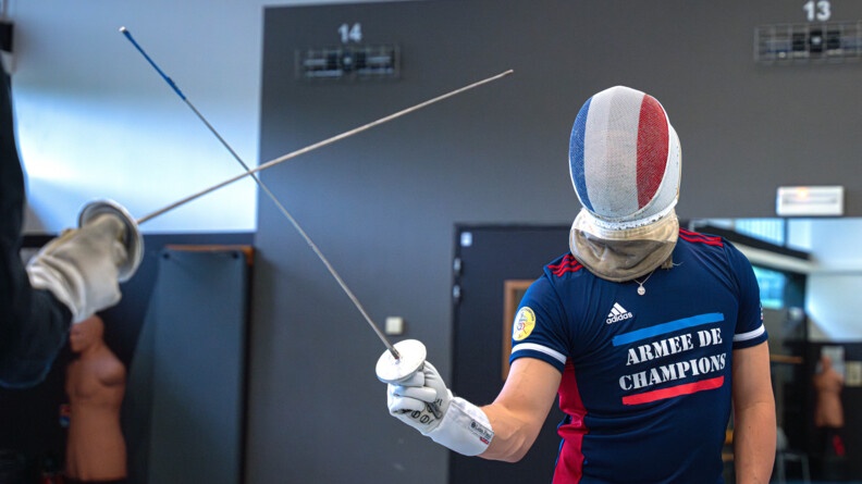 Maxime Pauty vêtu du survêtement bleu et rouge de l'armée de champions s'entraîne face à son maître d'arme dont on voi seulement le fleuret à gauche