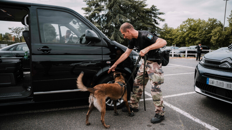 Agrandir l'image Un maître de chien gendarmerie en haut noir et bas de treillis montre la roue d'un véhicule grande capacité noir sur un parking