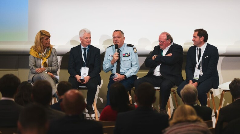 Au premier plan flou, toris rangs de personnes assistant à la table ronde. Au deuxième plan, de gauche à droite sur des tabourets hauts: Dominique Faure, un élu, le DGGN (le général d'Armée Christian Rodriguez) s'exprimant au micro, et deux autres élus.