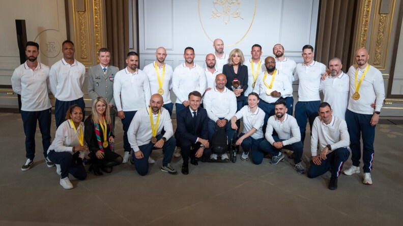 Dans une salle de l'Elysée, un groupe de militaires et de gendarmes en pantalons de survêtement bleus et polos manches longues blancs entourent le président de la République, agenouillé à côté d'un militaire en fauteuil roulant, derrière lequel se tient Madame Brigitte Macron.