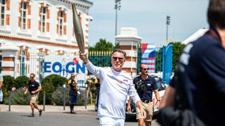 Un homme vétu de blanc élève la flame olympique, de ârt et d'autre trois hommes vêtus d'un polo bleu et short beige. En fond la grille de l'Ecole des officiers de la gendarmerie nationale