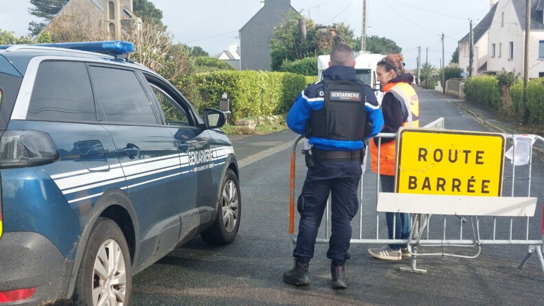 Sur la gauche de la photo, apparait la voiture bleue de la gendarmerie. Au centre de la photo, un gendarme se tient debout, de dos, et discute avec une femme vêtue d'un gilet orange de sécurité. Entre eux, se dresse une barrière sur laquelle il est inscrit Route barrée. Derrière eux, on aperçoit la rue bordée de maisons.