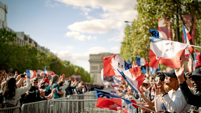 La foule en liesse de part et d'autre de barri&egrave;res, agitant des drapeaux fran&ccedil;ais. Dans le fond, l'Arc de Triomphe.