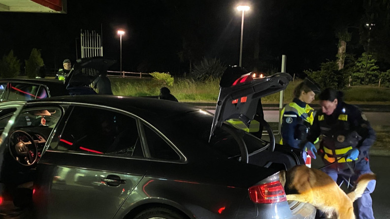 De nuit en ext&eacute;rieur, deux gendarmes dont un maitre chien et chien sont en train de fouiller le coffre d'une voiture.